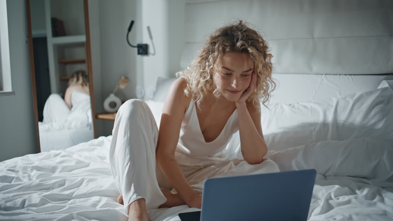 Menina feliz navegando no laptop na cama. Mulher despreocupada acessando a internet na luz da manhã.