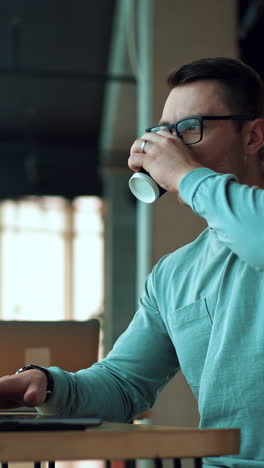 man working on laptop and drinking coffee from a paper cup. positive smiling businessman typing on computer sitting in comfortable office. vertical video