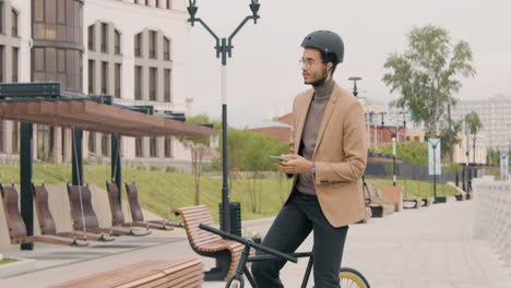 young american man in formal clothes and helmet using mobile phone while sitting on bike in the city