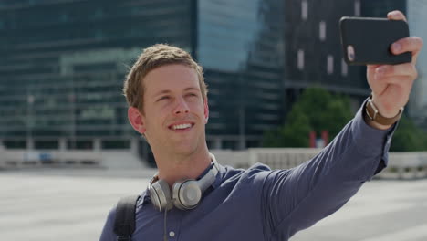 Retrato-De-Un-Joven-Atractivo-Usando-Un-Teléfono-Inteligente-Sonriendo-Tomando-Una-Foto-Selfie-En-La-Ciudad-Disfrutando-Compartiendo-Experiencias-De-Viaje-Urbano-En-La-Tecnología-De-La-Cámara-Del-Teléfono-Móvil