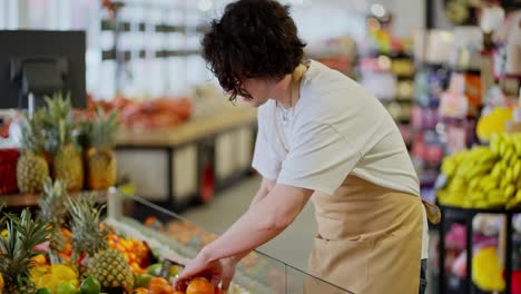 A-confident-guy-a-brunette-store-worker-with-curly-hair-in-a-white-T-shirt-and-a-yellow-apron-lays-out-goods-on-the-counter-in-a-supermarket