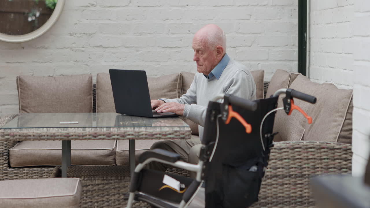 Senior man using laptop at home with wheelchair nearby