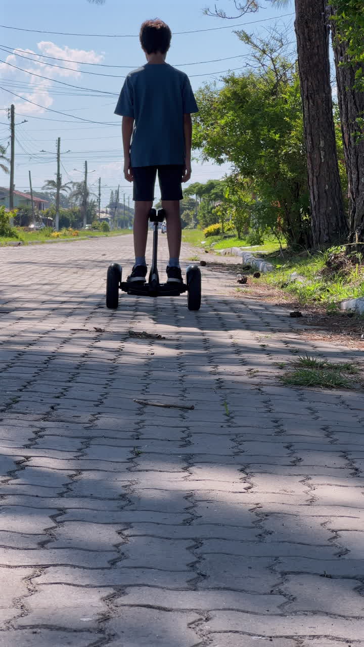Child rides hoverboard away from camera along residential street, vertical video with trees and power lines, brick pavement in daylight, 4K30