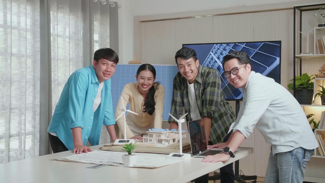 아시아 엔지니어 그룹은 사무실에서 태양 전지판을 가진 작은 집 모델에 대해 일하면서 카메라에 미소 짓고 있습니다.