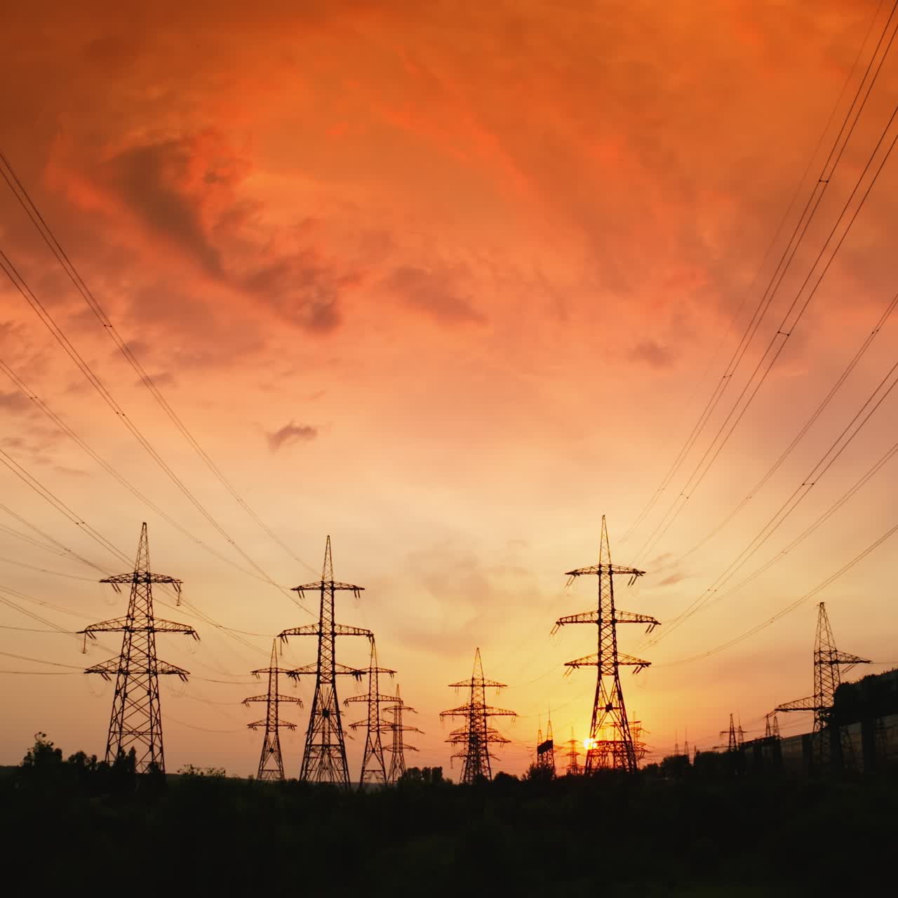 High voltage cable and pylons at sunset. Silhouettes of electric towers against dramatic sky with red setting sun