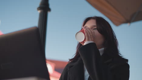 woman in black coat sipping juice, laptop in front, drops cup, continues working, background features blurred red roof and decorative pole