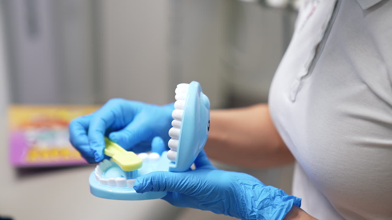 Female medic in blue latex gloves holding a denture and toothbrush. Dentistry specialist is showing how to clean teeth correctly. Close up.