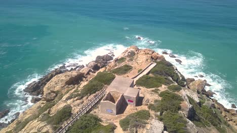 Toma-Aérea-De-Un-Cabo-En-El-Sur-De-España-Con-Las-Olas-Golpeando-Las-Rocas-Y-Una-Pasarela-De-Madera