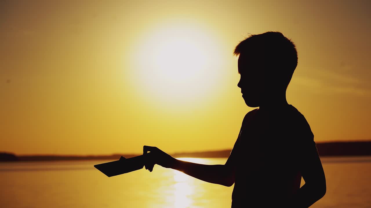 boy is making origami in the form of a ship on the background of sunset by the river in the summer