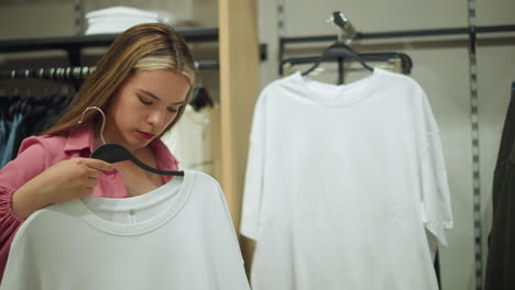 young lady wearing an unbuttoned pink top holds a white shirt against her body, thoughtfully evaluating it, while other clothes, including ash trousers, are neatly hanged on the racks