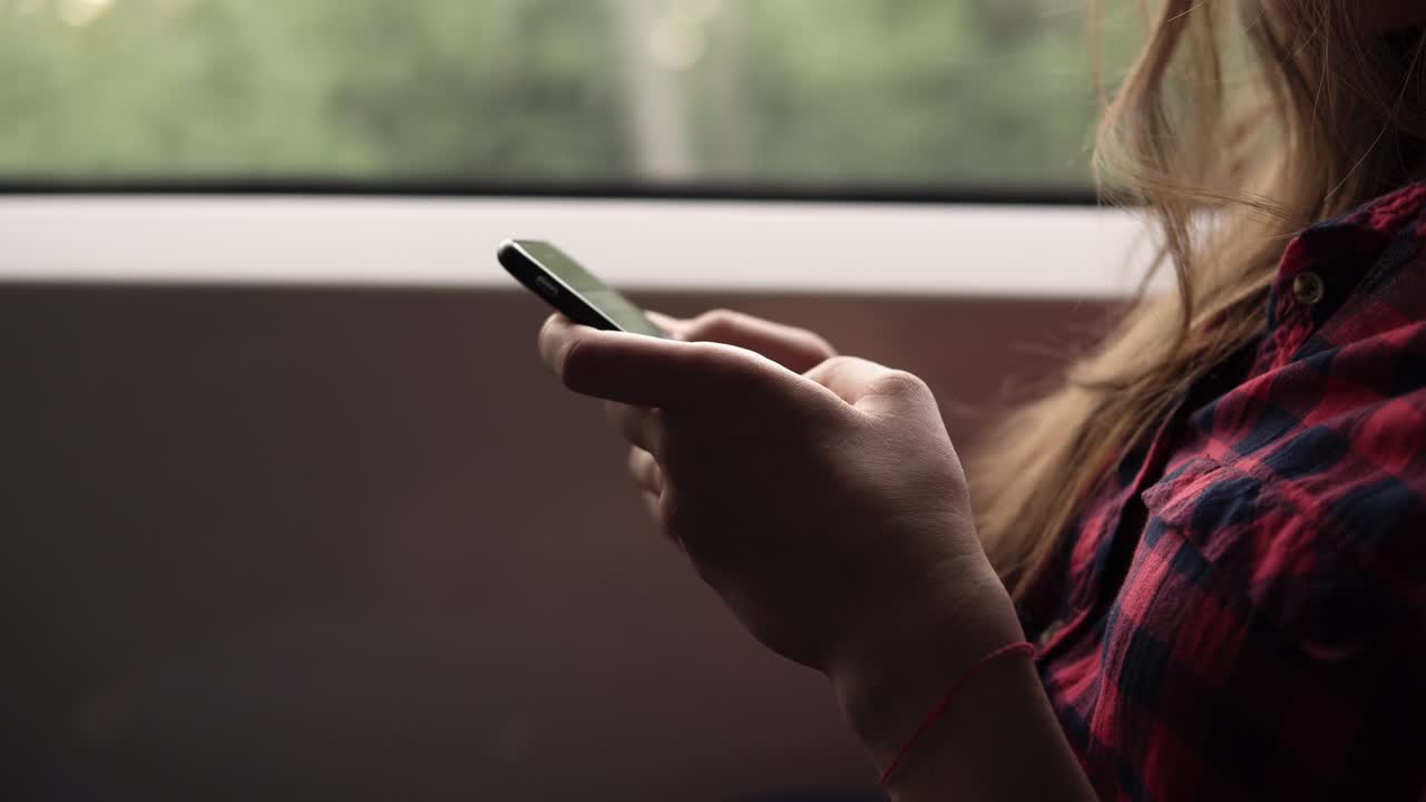 Extremely close up of woman's hands with mobile phone. Casual plaid shirt. Pretty girl is travelling by train. Nature landscape in window in motion. Side view