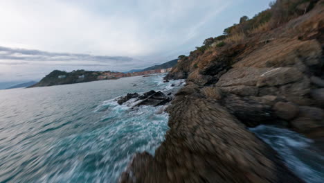 FPV-drone-flies-over-rocky-coastline-with-blue-ocean-and-distant-town