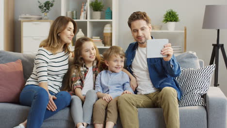 familia con dos niños sentados en el sofá y agitando las manos mientras tienen videochat en la tableta