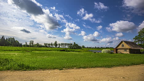 Timelapse-shot-of-a-man-mowing-the-grass-in-front-of-a-house-beside-a-lake-with-white-clouds-passing-at-daytime