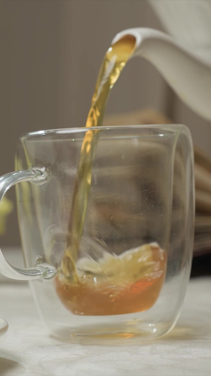 Una mano si allunga verso una teiera bianca per versare del liquido in una tazza di vetro. Il tè entra costantemente e il livello sale quasi fino all'orlo. Una superficie bianca testurizzata sostiene il bicchiere mentre la teiera è sfocata.