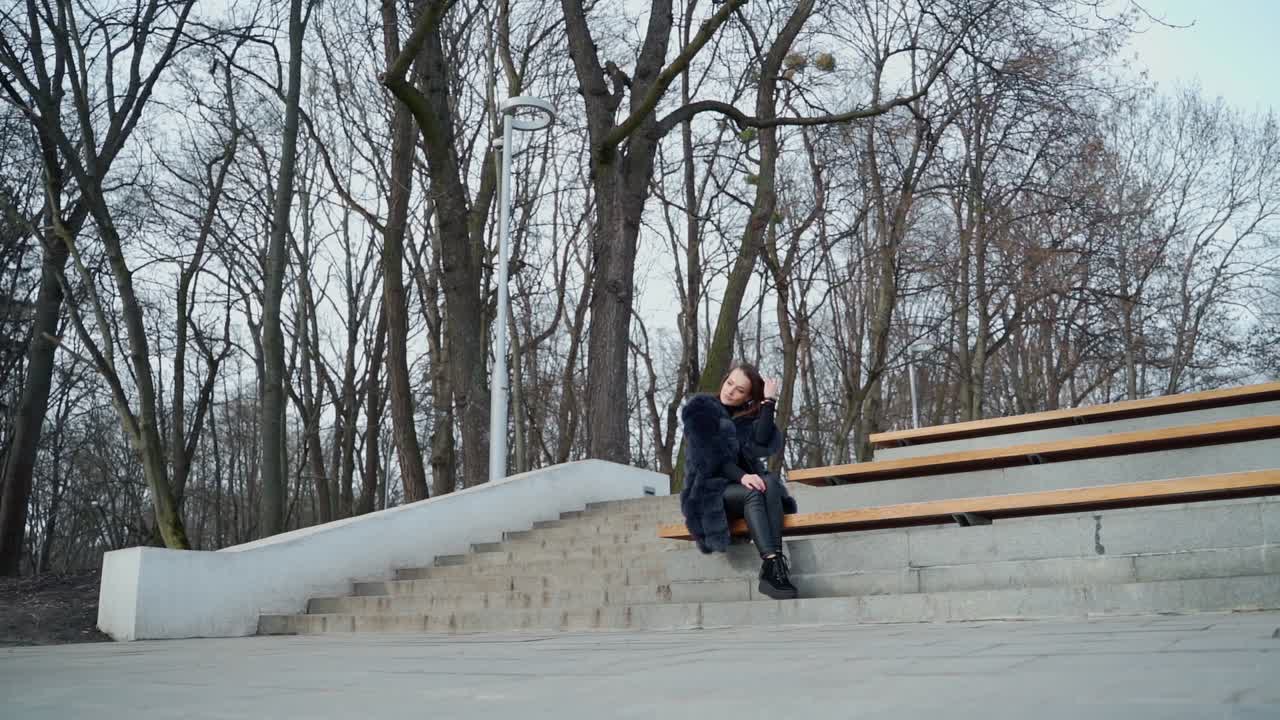 Girl sits on bench in the park. Beautiful young woman in fluffy coat sitting alone outdoors on the leafless trees background. View from below.