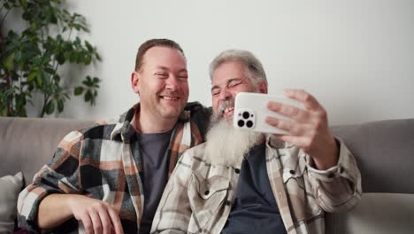 Happy-gay-couple-a-middle-aged-man-with-gray-hair-and-a-lush-beard-takes-a-selfie-using-a-White-phone-along-with-his-brunette-boyfriend-in-a-plaid-shirt-while-sitting-on-the-sofa-in-a-modern-apartment