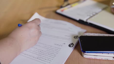 a close up of a female hand that writing on a paper on the table, a mobile, notebook, pen are on the table