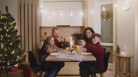 Blonder-Junge,-Der-Mit-Seiner-Schwester-Und-Seinen-Eltern-Am-Weihnachtstisch-Sitzt,-Benutzt-Ein-Smartphone,-Um-Ein-Familienselfie-Zu-Machen-1