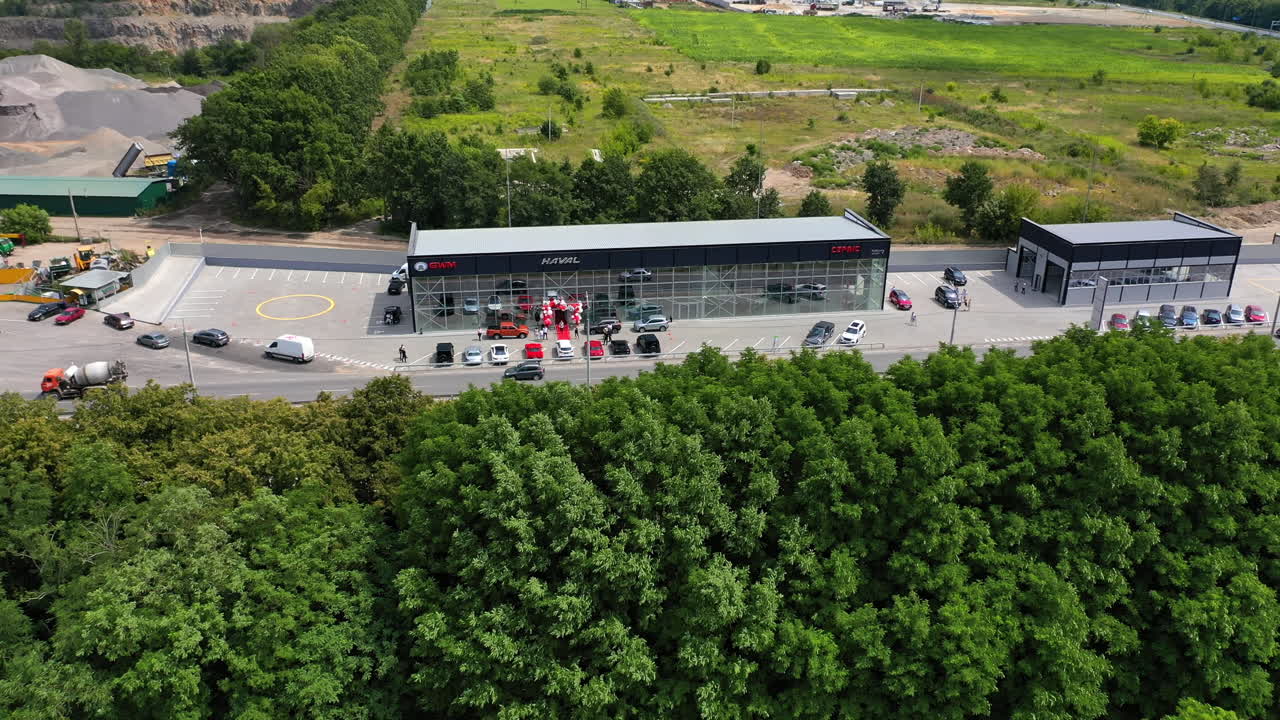 Transport goes by the road passing through the newly-opened car dealership. Drone distancing and rising over the green trees.