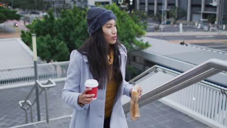 Asian-woman-walking-holding-takeaway-coffee-and-eating-sandwich