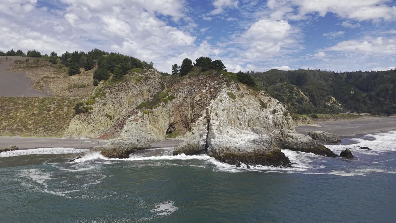 Un drone scivola lateralmente lungo una scogliera rocciosa mentre le onde si infrangono su una spiaggia di sabbia scura con colline boscose vicino a Constitución, Maule Cile, 4K.