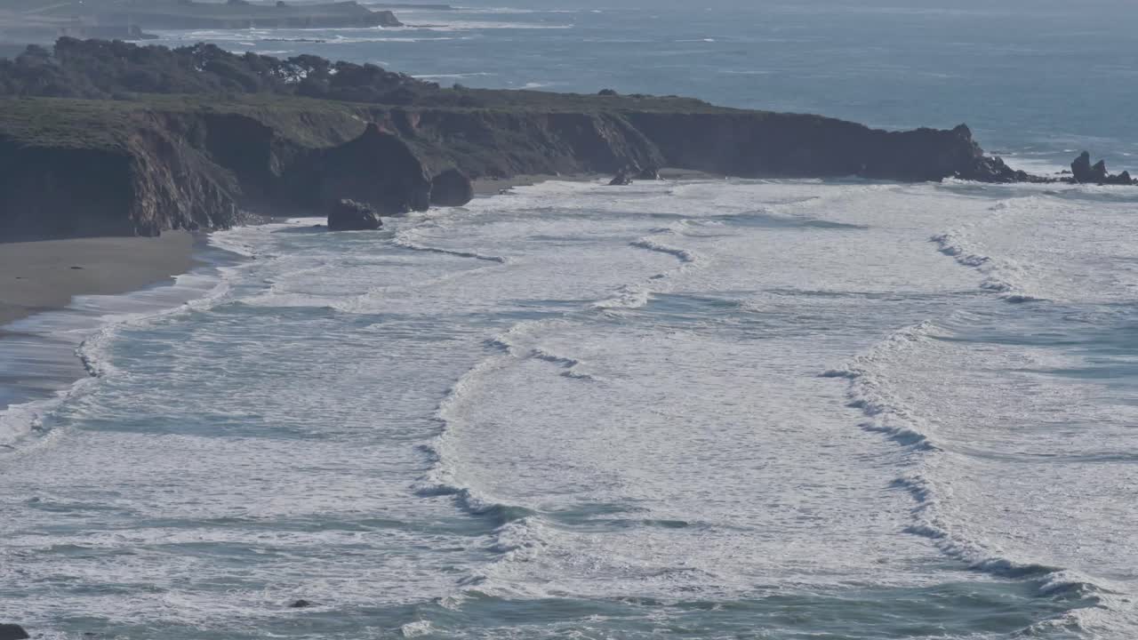 Aerial view of waves breaking in rhythmic curves along the rugged Big Sur coast, highlighting the natural power and beauty of California’s shoreline.