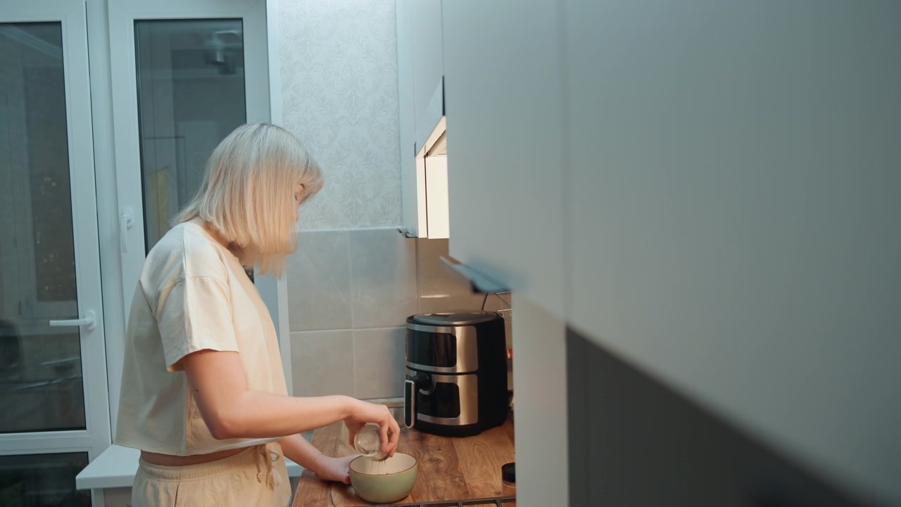 Blonde caucasian woman stirring bowl in cozy kitchen at night, warm undercabinet light, coffee maker on countertop, focused slow movements, casual shirt, textured tile backdrop, domestic calm