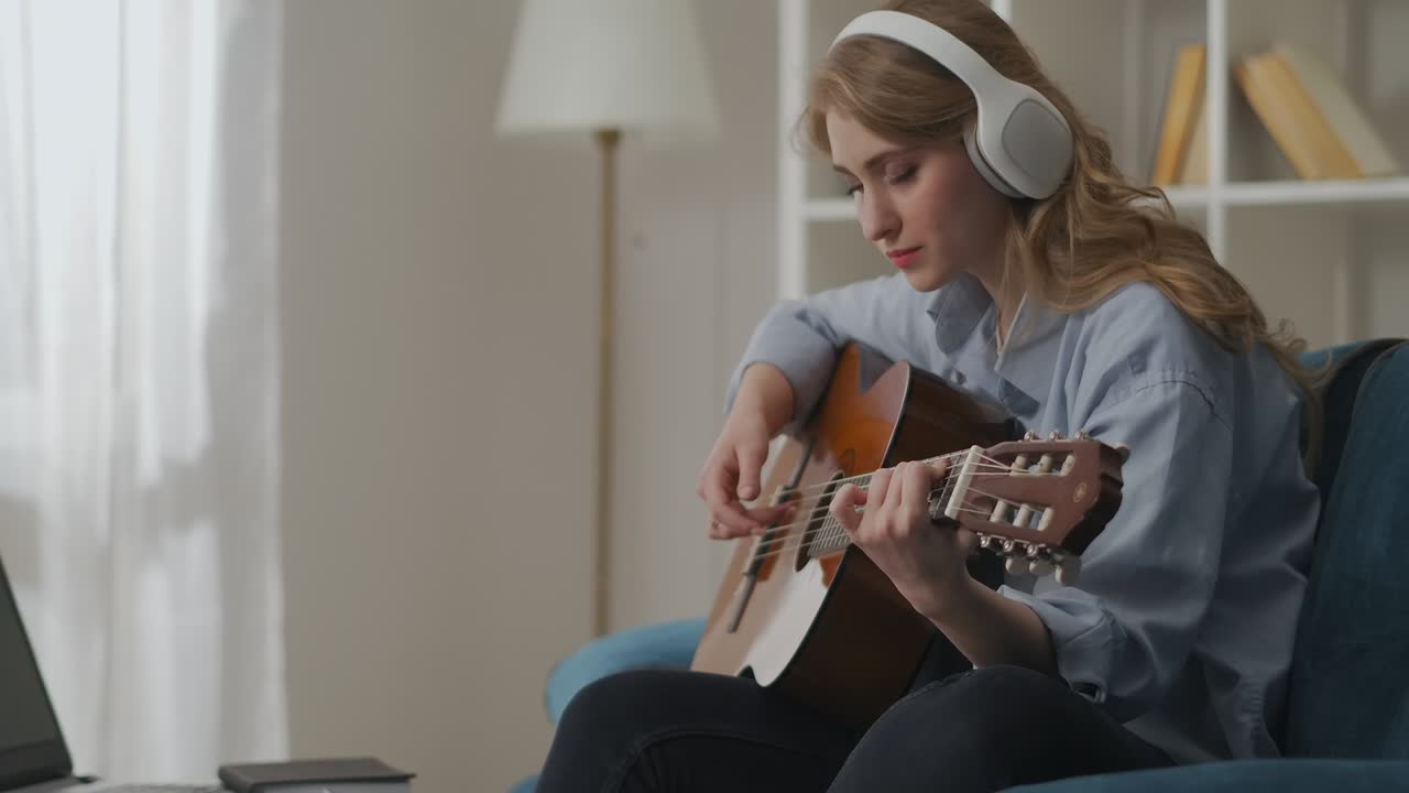 young woman is playing melody on guitar and swaying head in beat, sitting alone on couch in living room