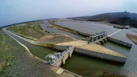 Top-Aerial-view-of-spillway-of-a-dam,-Beautiful-majestic-mountains-in-the-back-of-the-dam,-Sun-shines-in-the-green-water-of-the-dam