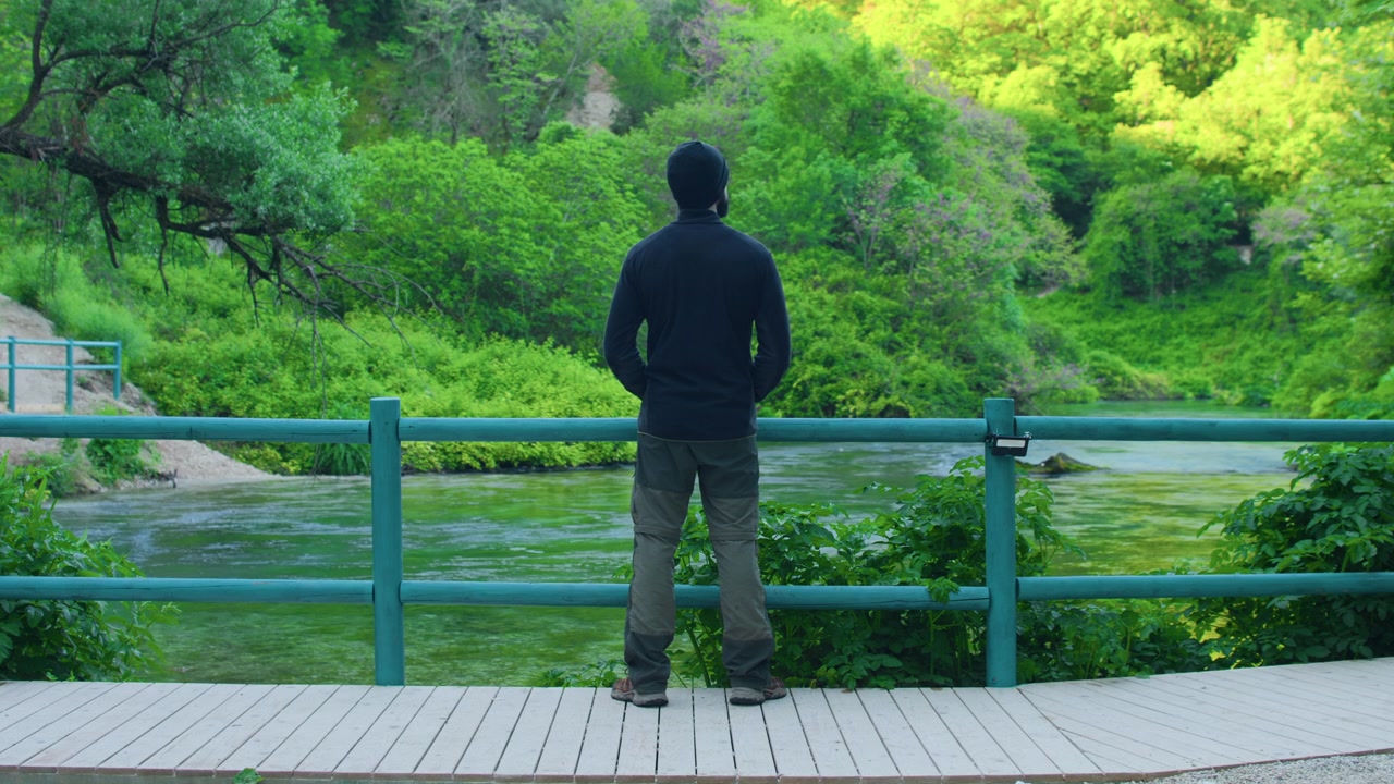 緑豊かな森の中で山の川を眺めるハイカーの男性、静止したワイドショット。木のフェンスのそばに立ち、透明な小川を眺める旅行者の後ろ姿。