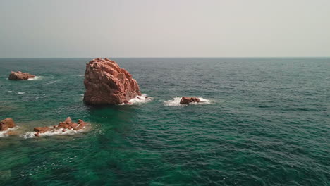 Big-Red-Rocks-With-White-Waves-Crashing-In-The-Ocean-Of-Sardinia,-Italy-At-Daytime---aerial-drone-shot