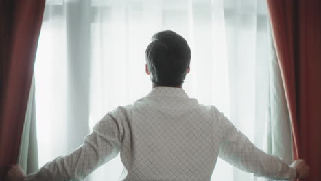 Back-view-of-young-man-opening-curtains-Closeup-handsome-guy-standing-by-window.