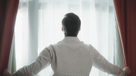 back view of young man opening curtains closeup handsome guy standing by window.