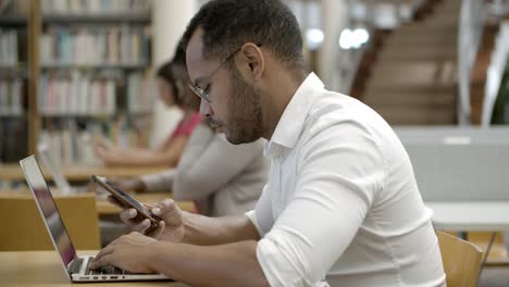 Vista-Lateral-Del-Joven-Enfocado-Usando-Un-Teléfono-Inteligente-En-La-Biblioteca