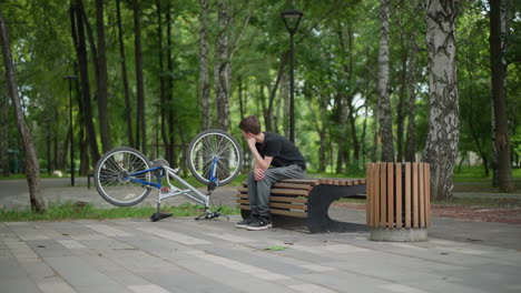 young boy in black top sits thoughtfully on park bench, resting his chin on his hand beside his upside-down bicycle, a black bird flies slightly blurred in the background