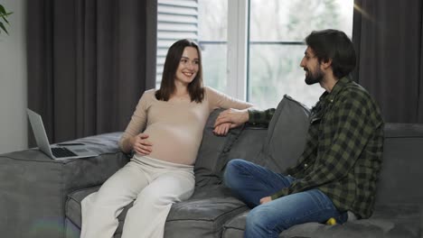 Woman-touching-pregnant-belly,-sitting-with-friend-or-husband-on-sofa