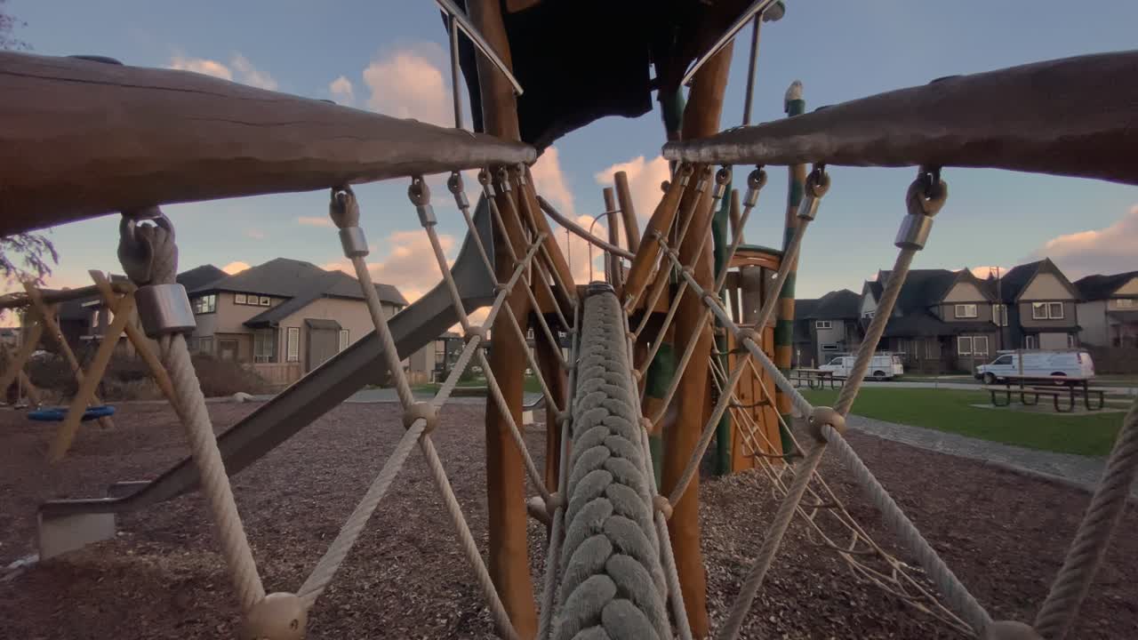 Thick woven climbing rope stretching across timber play structure hanging above wood chips framed against suburban housing beneath cloudy sunset sky captured via backward moving viewpoint