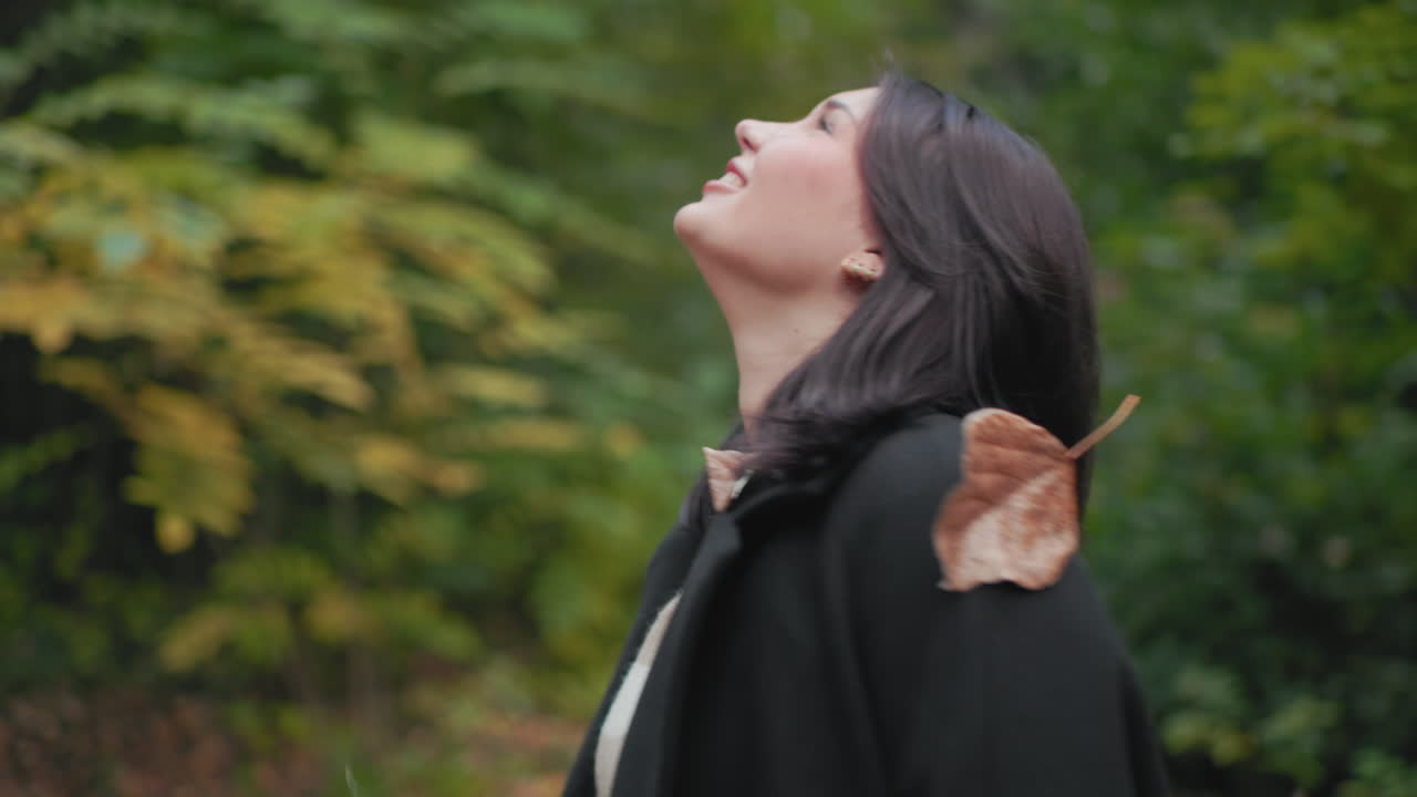 검은색 코트를 입은 우아한 여성이 마른 나뭇잎을 들고 나뭇잎을 던져 올린 다음, 초록빛 가을 숲에서 밝게 웃으며 빙글빙글 돕니다. 장난기 넘치고 활기찬 분위기에서 편안한 산책을 하며 솔직한 계절의 즐거움을 만끽해요.