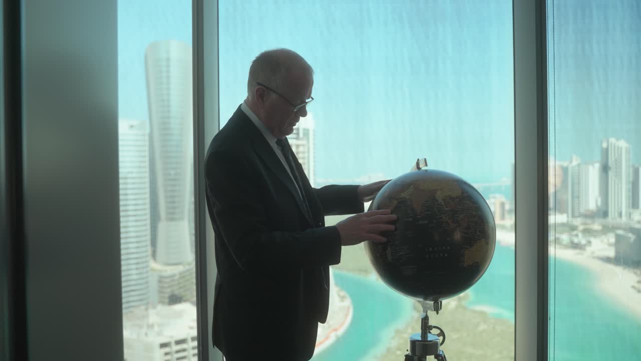 Senior businessman in a high-rise office with a stunning view of the city uses a globe as he considers his next business plan - slow motion