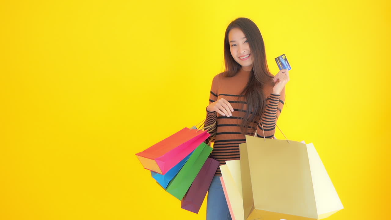 Slow-motion of Attractive Asian girl holding many shopping bags and showing credit plastic card on a yellow studio background, excited face expression