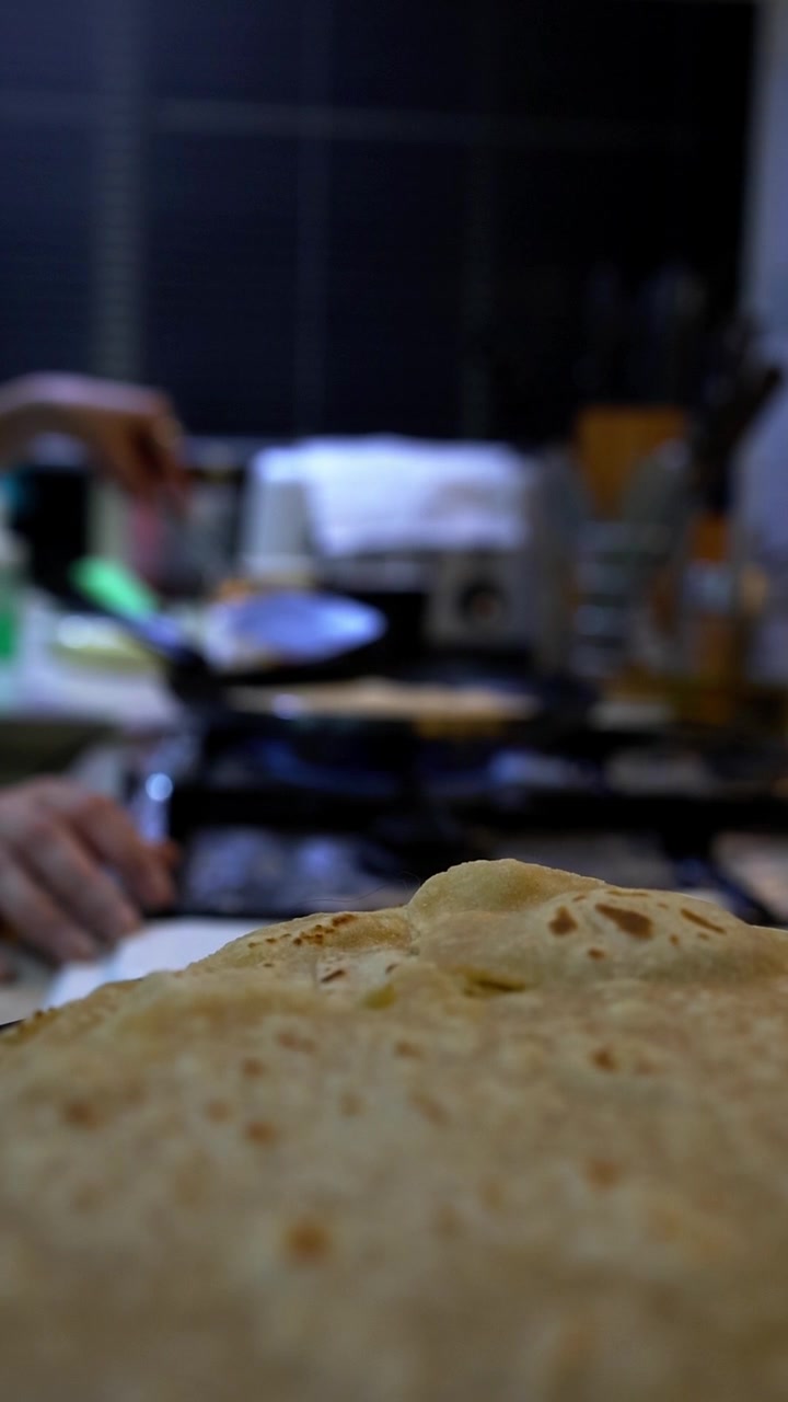 Close-up of freshly made chapati flatbread with visible bubbles and char marks. Woman's hands work at a kitchen counter with cooking utensils and equipment blurred in background. Warm, golden-brown br