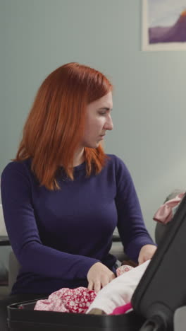 pensive woman with red hair puts clothes into open suitcase sitting on couch near bright lamp. housewife feels sad before going to vacation slow motion