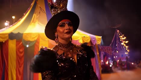 a woman in a black costume with a large hat, standing in front of a circus tent at night, is performing on stage.