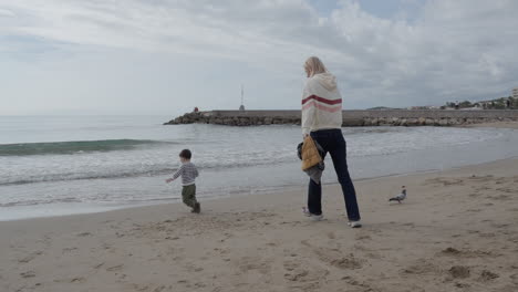 Toddler happily runs around sea side chasing pigeons as his mother runs after him