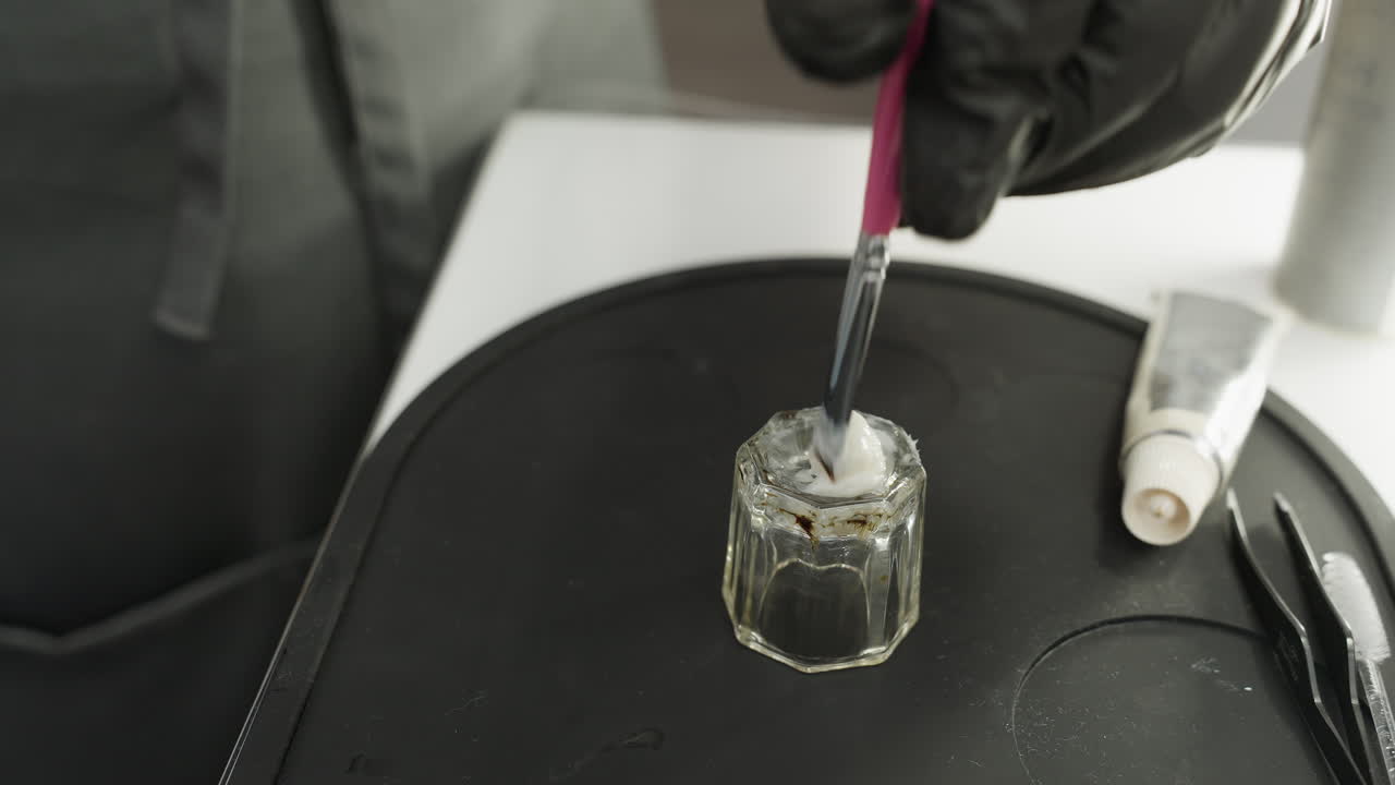 Close-up of technician wearing black gloves using pink brush to mix white cream inside transparent glass jar placed on black silicone tray with beauty tools and squeezed cosmetic tube nearby
