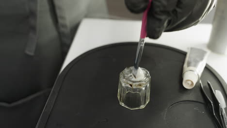 close-up of technician wearing black gloves using pink brush to mix white cream inside transparent glass jar placed on black silicone tray with beauty tools and squeezed cosmetic tube nearby