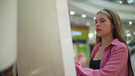 lady in pink dress intensely focused on her task, using a screen in a brightly lit mall, people can be seen in the background on an escalator, with blurred lights and other items in the background