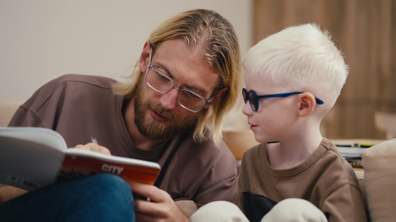 Close-up shot of a small albino boy with white hair in round blue glasses together with his dad, a blond man in glasses with a beard, doing his homework and teaching him to write and read in a modern kitchen in the evening at home