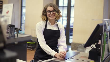 Woman-in-black-apron-as-a-cashier-at-the-cash-register-in-the-supermarket-or-discounter.-Smiling-short-haired-curly-woman-smiling-to-the-camera